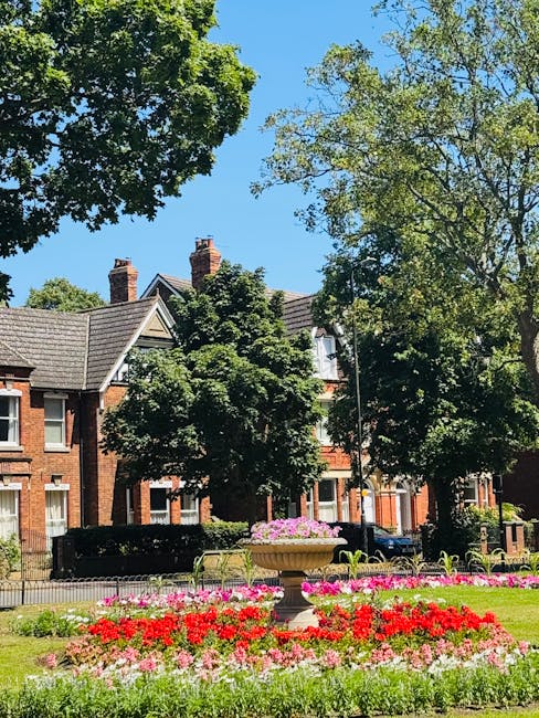 A well-maintained urban garden scene featuring a neatly trimmed lawn with lush green grass, bordered by vibrant flower beds filled with pink, red, and purple blooms. There is a large stone planter in